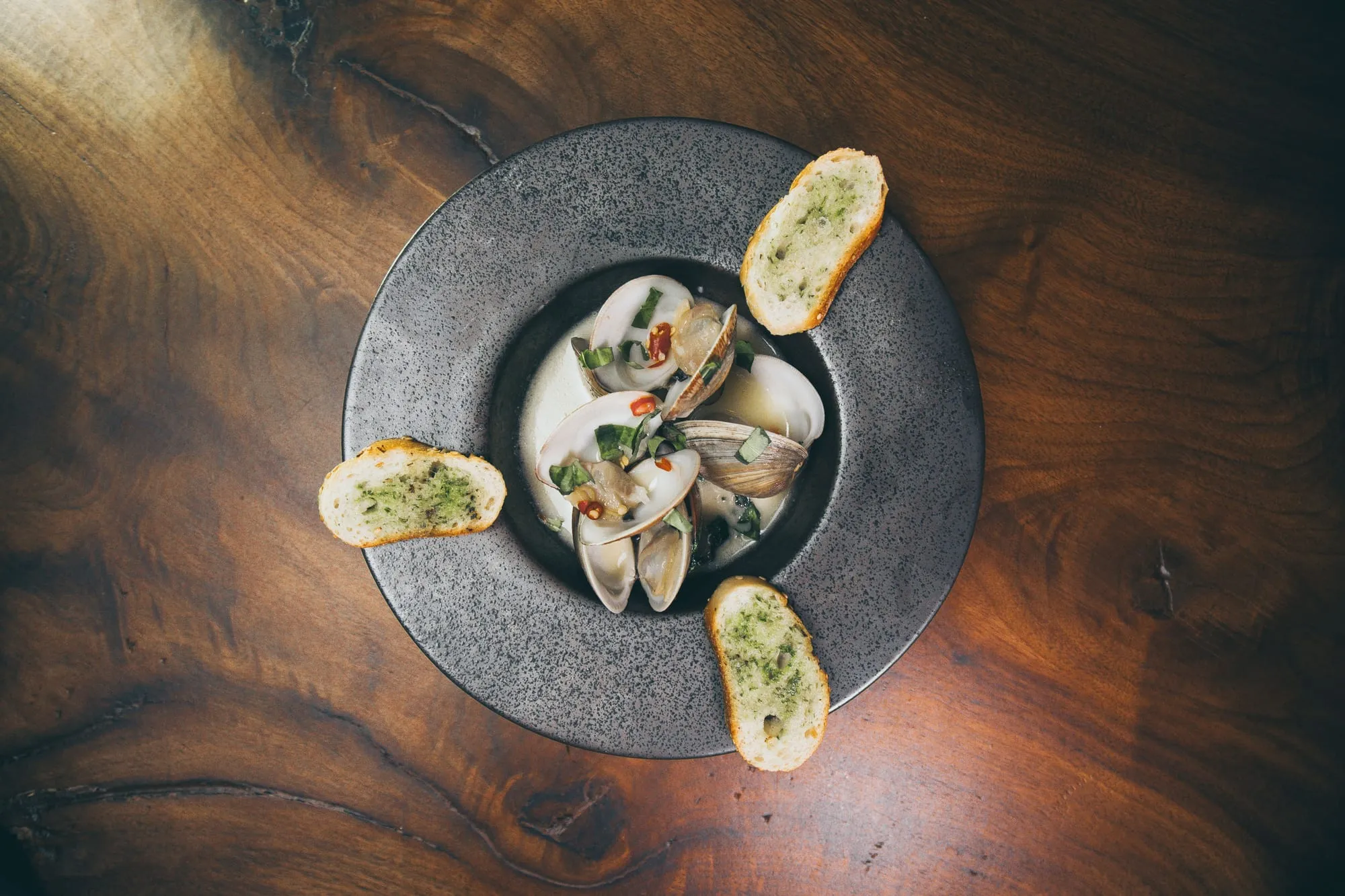 Clams in broth on a dark ceramic plate with slices of herbed garlic bread.