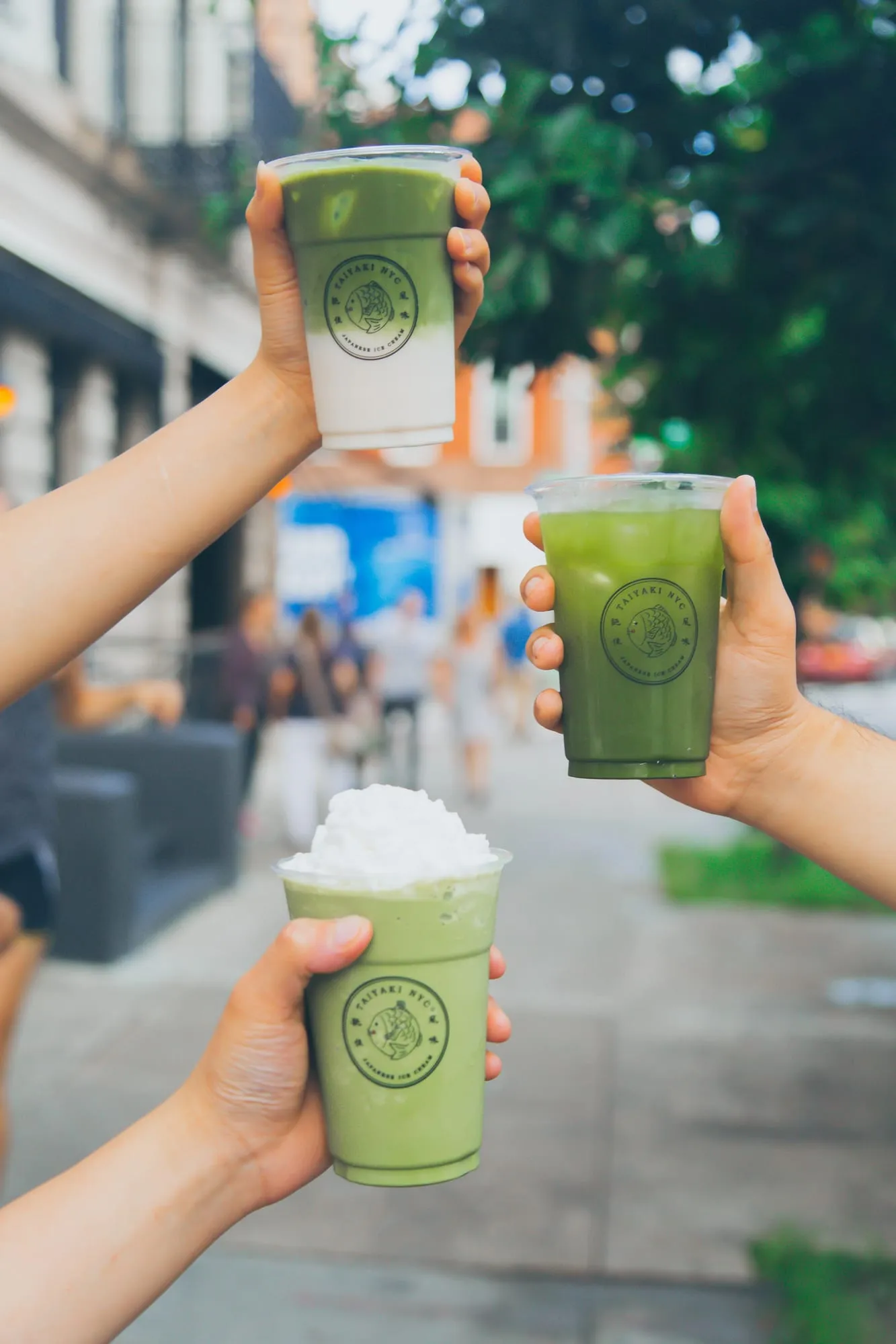 Two hands raising iced matcha drinks toward each other on a city street.