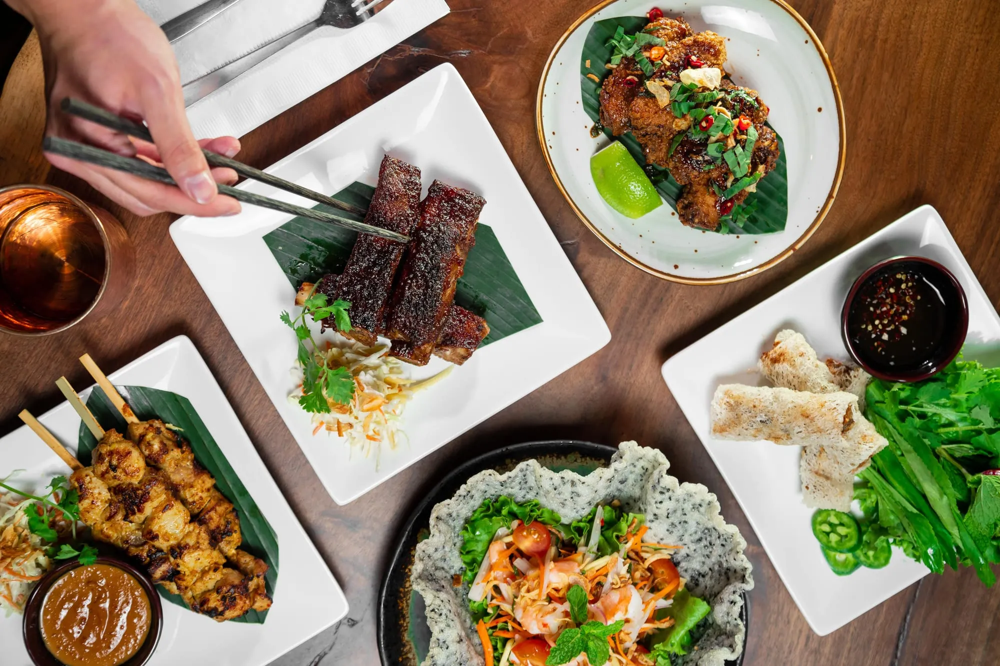 Chopsticks lifting a skewer above several plated restaurant dishes on a wooden table.