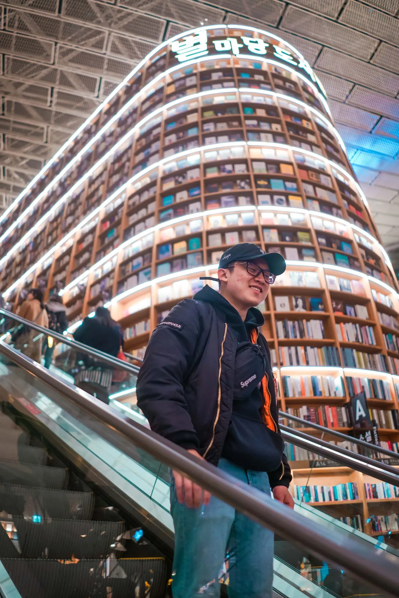 A man on an escalator inside a bookstore with towering curved shelves behind him.