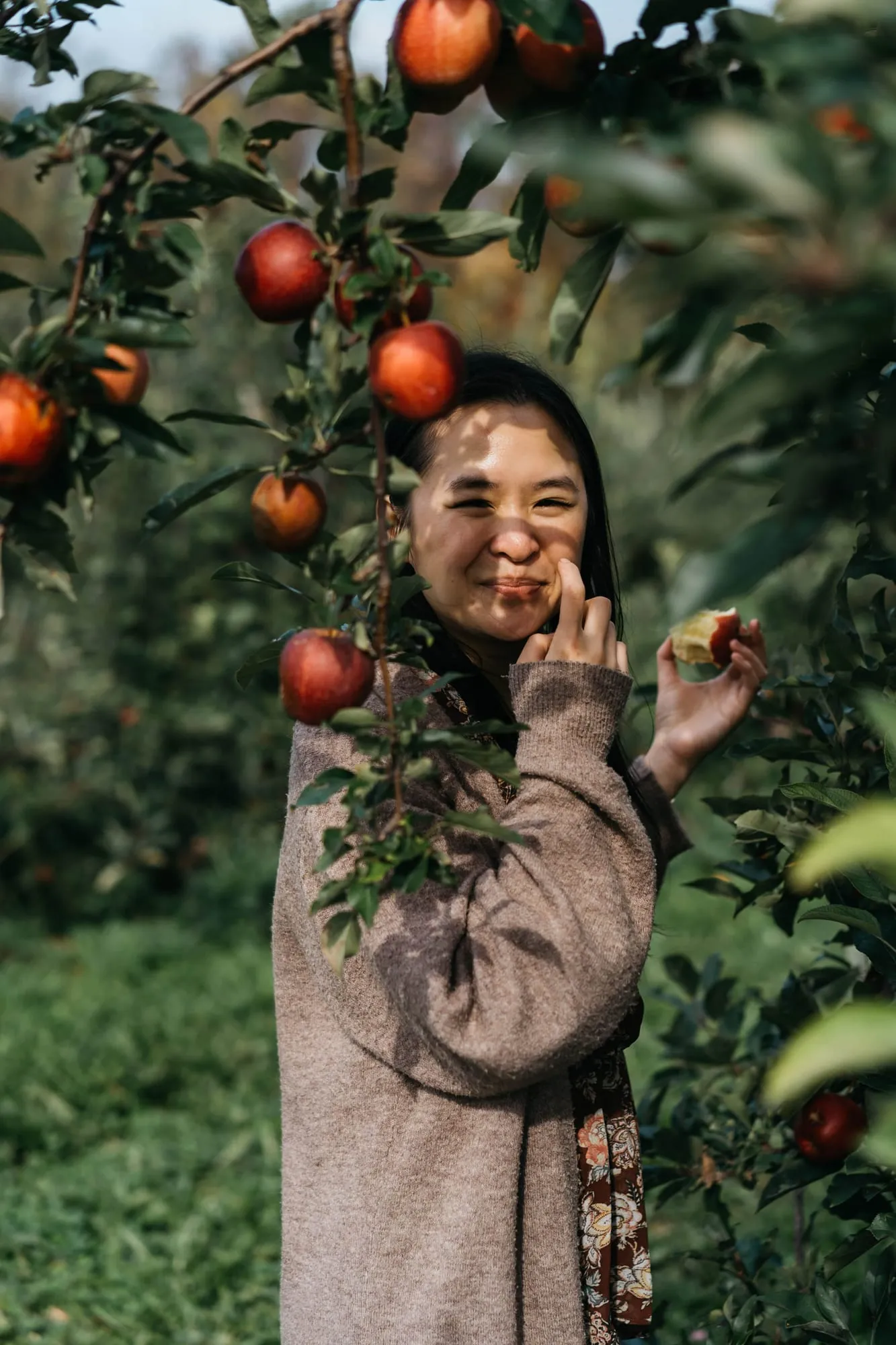A woman smiling beside a pomegranate tree, reaching up toward the fruit.